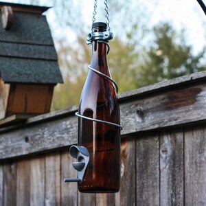 K&ouml;nnte beinhalten: Eine braune Glasflasche mit einem Metalldrahtk&auml;fig und einem daran befestigten Metallfutterspender. Die Flasche h&auml;ngt an einer Kette, die an einem schwarzen Metallhaken befestigt ist. Die Flasche h&auml;ngt vor einem Holzzaun.