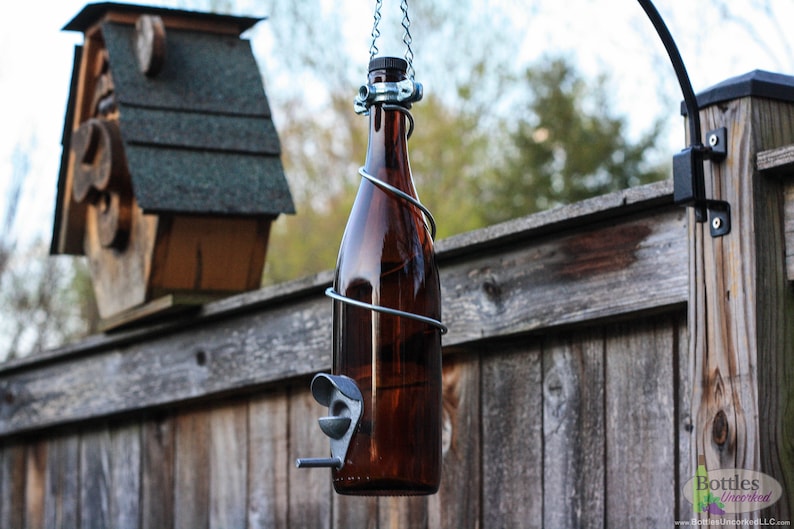 K&ouml;nnte beinhalten: Eine braune Glasflasche mit einem Metalldrahtk&auml;fig und einem daran befestigten Metallfutterspender. Die Flasche h&auml;ngt an einer Kette, die an einem schwarzen Metallhaken befestigt ist. Die Flasche h&auml;ngt vor einem Holzzaun.