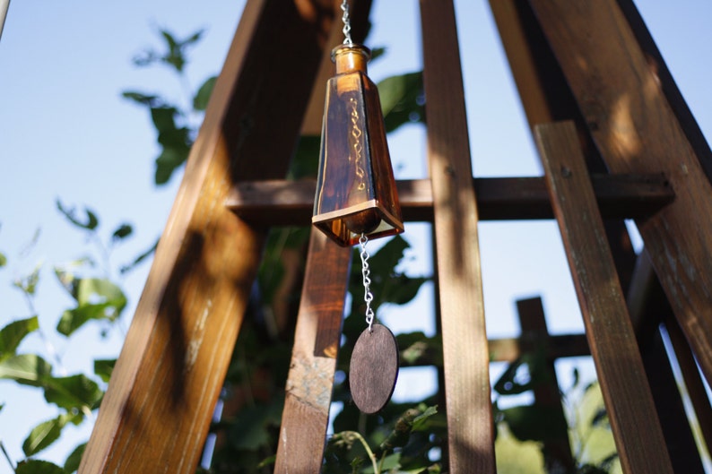 Puede incluir: Un carill&oacute;n de viento de botella de vidrio marr&oacute;n que cuelga de una estructura de madera. La botella tiene una cadena unida a ella con una pieza de madera redonda en el extremo.