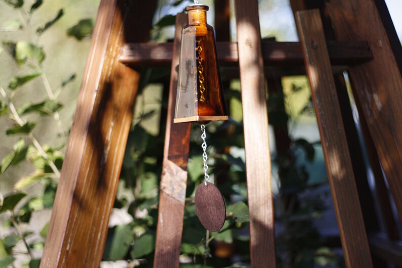 Puede incluir: Un carill&oacute;n de madera marr&oacute;n con una botella de vidrio y un disco redondo de madera colgando de una cadena plateada.