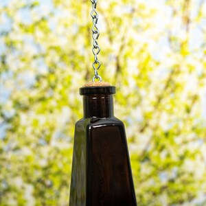 Puede incluir: Una botella de vidrio marr&oacute;n oscuro con un tap&oacute;n de corcho que cuelga de una cadena plateada. La botella est&aacute; suspendida frente a un fondo borroso de verde y blanco.