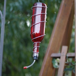 May include: A red glass bottle with a metal cage around it, used as a hummingbird feeder. The feeder is hanging from a metal hook.