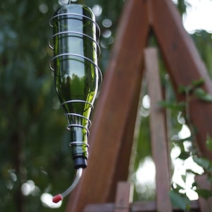 May include: A green glass bottle with a metal cage around it, hanging from a metal hook. The bottle has a red plastic spout at the bottom, and is likely a bird feeder.