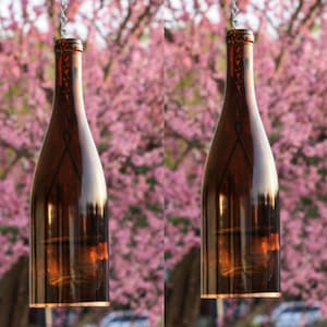May include: Two brown glass wine bottles hanging from chains against a blurred background of pink flowers. The bottles have a copper colored rim at the top and bottom.