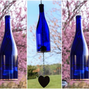 May include: Three blue glass bottles hanging from a chain with a heart-shaped charm at the bottom. The bottles are suspended in front of a blurred background of pink flowers and green trees.
