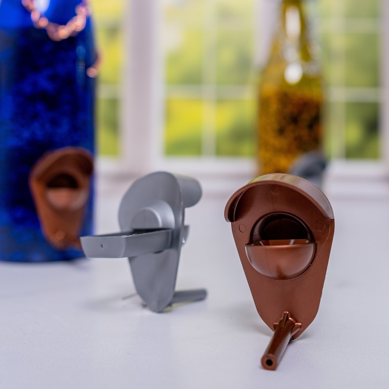 May include: Two bottle stoppers, one gray and one brown, with a spout and a small cup on top. The stoppers are on a white surface.