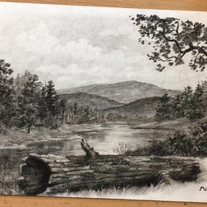 May include: A black and white charcoal drawing on paper depicting a serene landscape with a river flowing through a forest. A large fallen log rests in the foreground, while pine trees and rolling mountains fill the background under a cloudy sky.