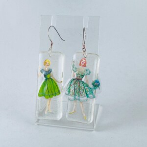 Puede incluir: Un par de pendientes colgantes con un dije rectangular de resina que presenta una ilustraci&oacute;n vintage de dos mujeres con vestidos. Una mujer lleva un vestido verde y la otra lleva un vestido de estampado floral.