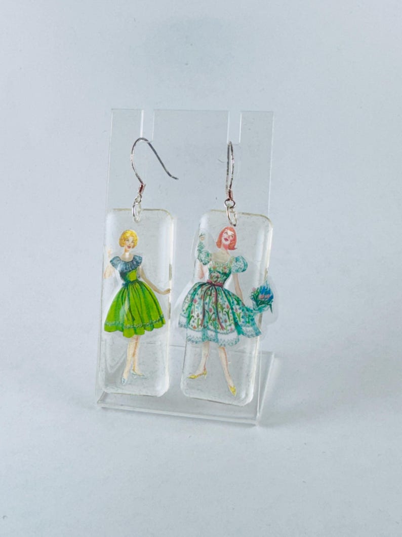 Puede incluir: Un par de pendientes colgantes con un dije rectangular de resina que presenta una ilustraci&oacute;n vintage de dos mujeres con vestidos. Una mujer lleva un vestido verde y la otra lleva un vestido de estampado floral.