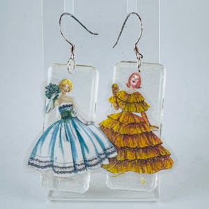 Puede incluir: Un par de pendientes de resina transparente con dos mujeres en vestidos vintage. Una mujer lleva un vestido azul y blanco con un ramo de flores, y la otra lleva un vestido amarillo con volantes.