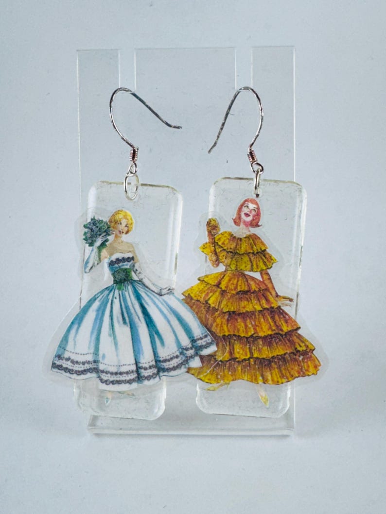 Puede incluir: Un par de pendientes de resina transparente con dos mujeres en vestidos vintage. Una mujer lleva un vestido azul y blanco con un ramo de flores, y la otra lleva un vestido amarillo con volantes.