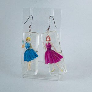 Puede incluir: Un par de pendientes colgantes con dijes rectangulares de resina transparente que presentan ilustraciones vintage de mujeres con vestidos. Una mujer lleva un vestido azul y la otra lleva un vestido rosa.
