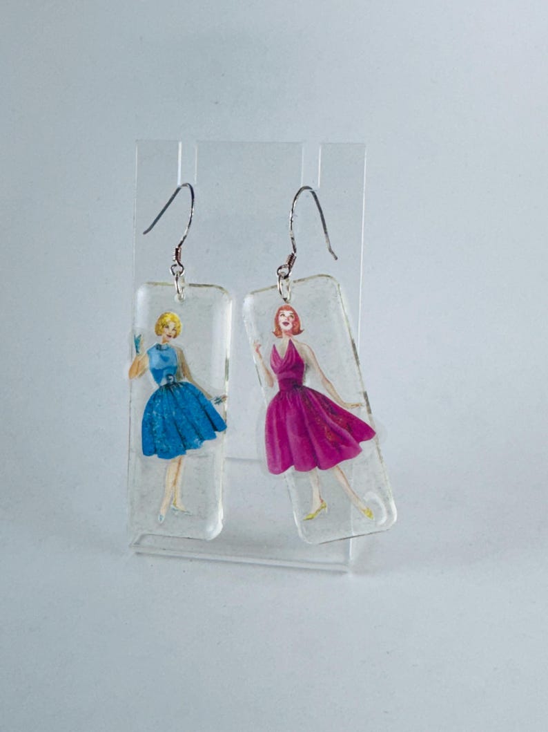 Puede incluir: Un par de pendientes colgantes con dijes rectangulares de resina transparente que presentan ilustraciones vintage de mujeres con vestidos. Una mujer lleva un vestido azul y la otra lleva un vestido rosa.