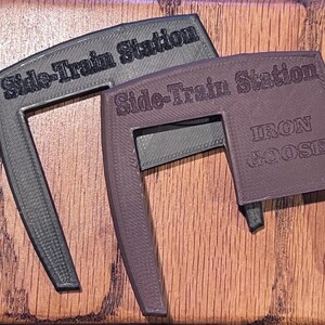 May include: Two 3D-printed, angled brackets. One is dark gray, the other is brown. Both have the text "Side-Train Station" and "Iron Goose" embossed on them. The brackets are on a wood-grain surface.