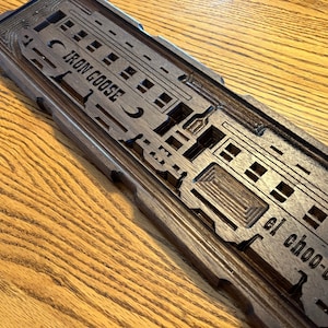 May include: A dark brown wooden sign with a carved design of a train. The sign reads "Iron Goose" and "El Choo-Choo".