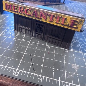 May include: A miniature model of a mercantile building with a yellow and red sign that reads "MERCANTILE". The building is dark brown with windows and a door. The model is on a green cutting mat.