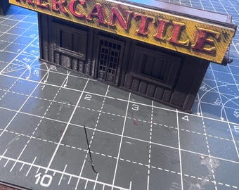 May include: A miniature model of a mercantile building with a yellow and red sign that reads "MERCANTILE". The building is dark brown with windows and a door. The model is on a green cutting mat.