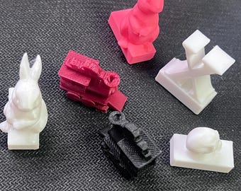May include: A collection of six small 3D printed objects including two white rabbits, one pink rabbit, one dark red train, one black train, one white letter X, and one white skull, all mounted on rectangular bases and arranged on a textured surface.