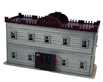 May include: A two-story miniature hotel building model with light gray horizontal siding, a dark maroon roof with a crenelated edge, and a central arched sign reading HOTEL. The structure features multiple windows with white curtains and a dark door.