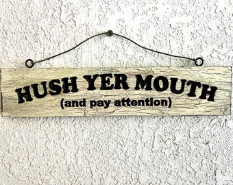 May include: A rectangular wooden sign with the text "HUSH YER MOUTH (and pay attention)" in black lettering. The sign has a distressed, off-white finish and is suspended by a curved wire.