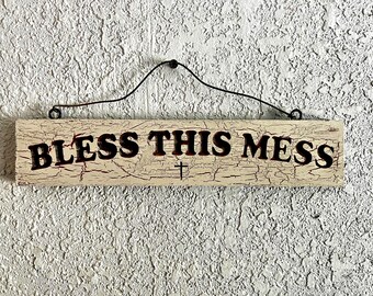 May include: A rectangular wooden sign with the words "BLESS THIS MESS" in dark brown letters. A small cross is below the text. The sign has a distressed, crackled finish and is suspended by a black wire.