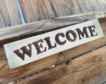 May include: A rectangular wooden sign with the word "WELCOME" in dark brown, raised letters. The sign has a white background with a light green border and is suspended by a wire. The sign is mounted on a weathered wooden fence.