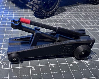 May include: A black 3D-printed model of a catapult with a red accent on the arm. The model has two small wheels and the initials "IG" are visible on the side. The catapult is on a gray surface with grid lines.