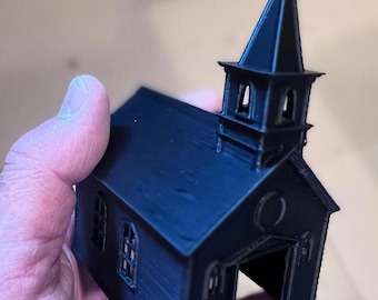 May include: A small, matte black 3D-printed model of a church building. The structure features a tall steeple topped with a cross, arched windows on the sides, and an open front doorway with a small rectangular platform extending from the entrance.