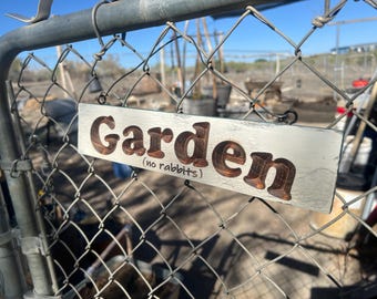 May include: A rectangular wooden sign with the word "Garden" in large, brown, raised letters. Below, in smaller text, it reads "(no rabbits)". The sign is attached to a chain-link fence.
