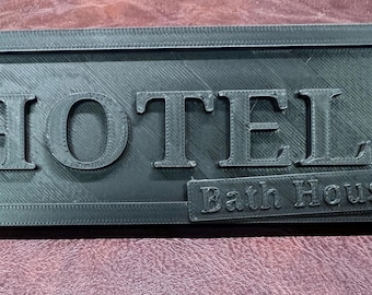 May include: A rectangular, dark gray sign with the word "HOTEL" in large, raised letters. Below, a smaller rectangle reads "Bath House". The sign has a textured surface and a framed border.