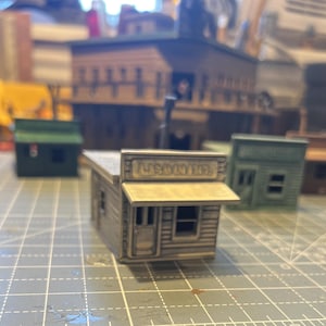 May include: A miniature model of a western-style building with a light brown awning and the word "SALOON" above the entrance. The building is gray with a wooden texture, and other miniature buildings are in the background.