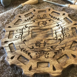 May include: A wooden serving tray with a decorative design of a floral wreath and a rectangular space in the center. The tray has a scalloped edge and 12 small rectangular slots around the perimeter.