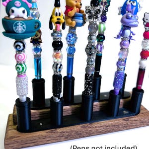 May include: A collection of decorative pens with various character toppers, including Winnie the Pooh, Hello Kitty, and a Starbucks-themed design. The pens are displayed in a wooden holder with black pen stands. The image text reads: (Pens not included).