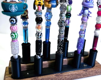 May include: A collection of decorative pens with various character toppers, including Winnie the Pooh, Hello Kitty, and a Starbucks-themed design. The pens are displayed in a wooden holder with black pen stands. The image text reads: (Pens not included).