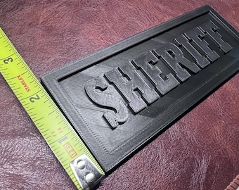 May include: Black rectangular sign with the word "SHERIFF" in raised, block letters. The sign is approximately 7 inches long and 3 inches wide. The sign is made of a textured material.