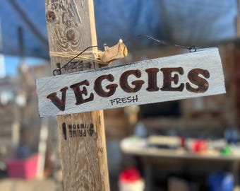 May include: A rectangular wooden sign with the word "VEGGIES" in dark brown letters and the word "FRESH" below it. The sign is hanging from a wooden post with a wire. The sign has a distressed, rustic look.