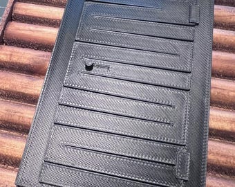 May include: A black, rectangular object with a textured surface and multiple parallel, raised lines. The object has a small, circular feature and is set against a wooden background. The design suggests a functional or architectural component.