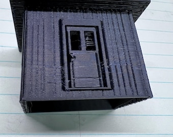 May include: A small, dark blue 3D-printed model of a miniature house or shed. The structure features a sloped roof, a front door, and a window. The walls have a textured, ridged appearance.