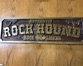 May include: A rectangular wooden sign with carved lettering that reads AUTHENTIC ROCK HOUND ROCK LICKER. The design features deep relief carving with dark brown stained recessed areas and lighter natural wood surfaces, including a pickaxe icon.