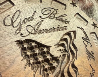 May include: A wooden plaque with the words "God Bless America" and an American flag design carved into the surface. The plaque features stars and decorative cutouts around the edges. The wood has a natural, light brown color.