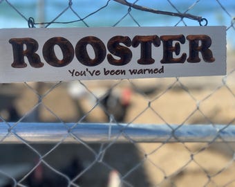 May include: A rectangular wooden sign with the word "ROOSTER" in large, brown, block letters. Below, the text reads "You've been warned." The sign hangs on a chain-link fence, with a blurred background of a chicken coop.