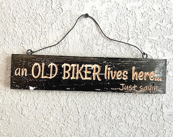 May include: Black wooden sign with the text "an OLD BIKER lives here... ...Just sayin" in a distressed, carved style. The sign is rectangular and hangs from a black wire. The background is a textured, off-white wall.