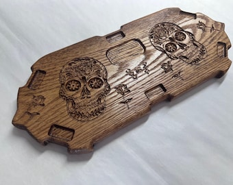 May include: A rectangular wooden board with a dark brown finish featuring laser-engraved designs of two ornate skulls and several roses. The board has a notched, decorative edge and a smooth, polished surface with visible wood grain patterns.