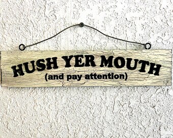 May include: A rectangular wooden sign with the words "HUSH YER MOUTH (and pay attention)" in black lettering. The sign has a distressed, crackled finish and is suspended by a curved metal wire.