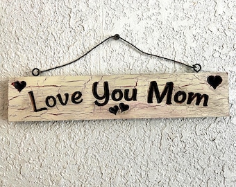 May include: A rectangular wooden sign with the words "Love You Mom" in black, featuring heart accents. The sign has a distressed, crackled finish with red undertones and is suspended by a black wire.