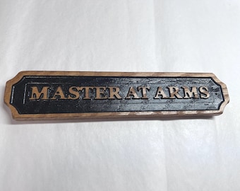 May include: A rectangular wooden sign with beveled edges featuring a black center panel. The text MASTER AT ARMS is embossed in a light wood tone across the center. The sign has a natural wood grain border surrounding the black recessed area.