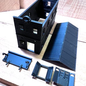 May include: A black, two-story model building with a star emblem, windows, and a separate roof. Additional components include a loading dock and doors, all in a dark color. The model is designed for hobbyists and model train enthusiasts.