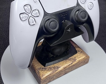 May include: A white and black gaming controller resting on a black stand with a wooden base. The controller has buttons with symbols and two black joysticks. The stand is on a white circular surface.