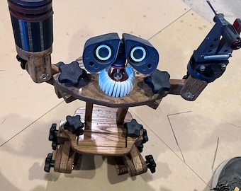 May include: A wooden robot-shaped tool holder featuring two articulated arms, one holding a cylindrical tool and the other a clamp. The robot has two dark blue eye-like components, a white gear-like mouth, and black star-shaped adjustment knobs.