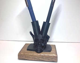 May include: A pen holder featuring a black skull design with two pen slots. The holder is mounted on a rectangular wooden base. Two pens, one blue and one dark gray, are placed in the slots.